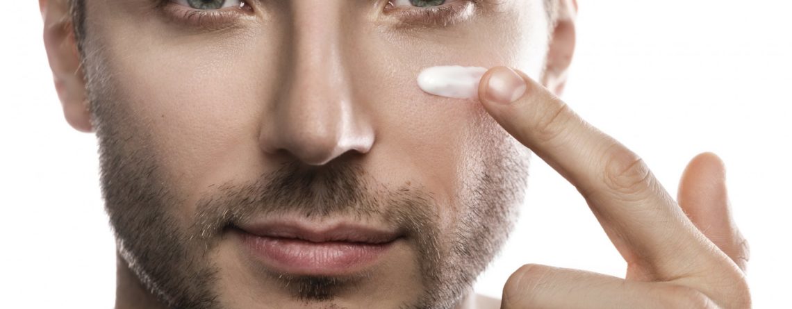 Ein Mann trägt eine Creme auf seine Wange auf, während er in die Kamera schaut. Hintergrund ist neutral.