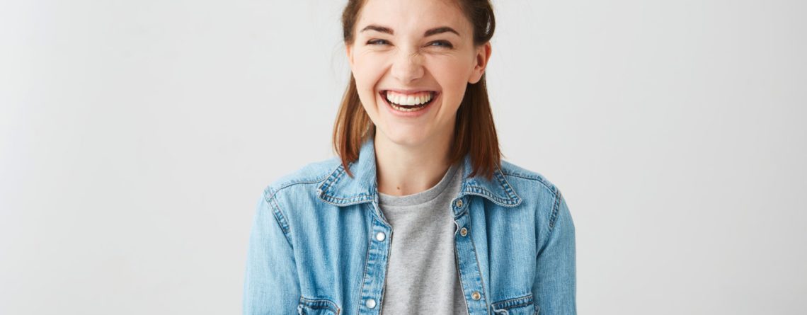 Lachende junge Frau mit Duttfrisur, trägt eine Jeansjacke über einem grauen T-Shirt, vor einem hellen Hintergrund.