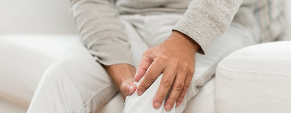 Mann sitzt auf einer Couch und hält sich schmerzerfüllt das Knie mit der Hand.