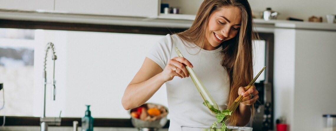 Frau mischt Salat in einer Schüssel in einer modernen Küche, umgeben von frischen Zutaten.