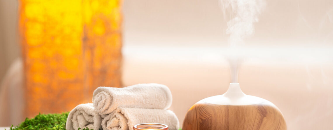 Ein Aromadiffusor aus Holz mit aufsteigendem Dampf, umgeben von Handtüchern, einer Kerze und grünem Moos.