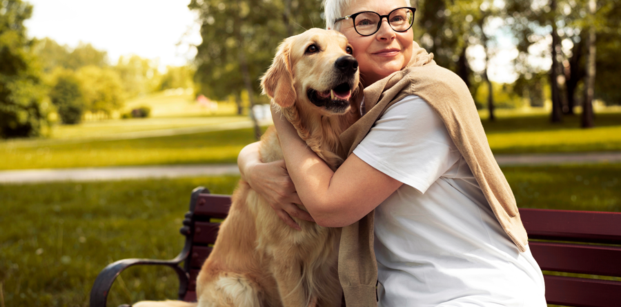 Eine Frau mit kurzen, blonden Haaren und Brille umarmt einen golden retriever auf einer Bank im Park.